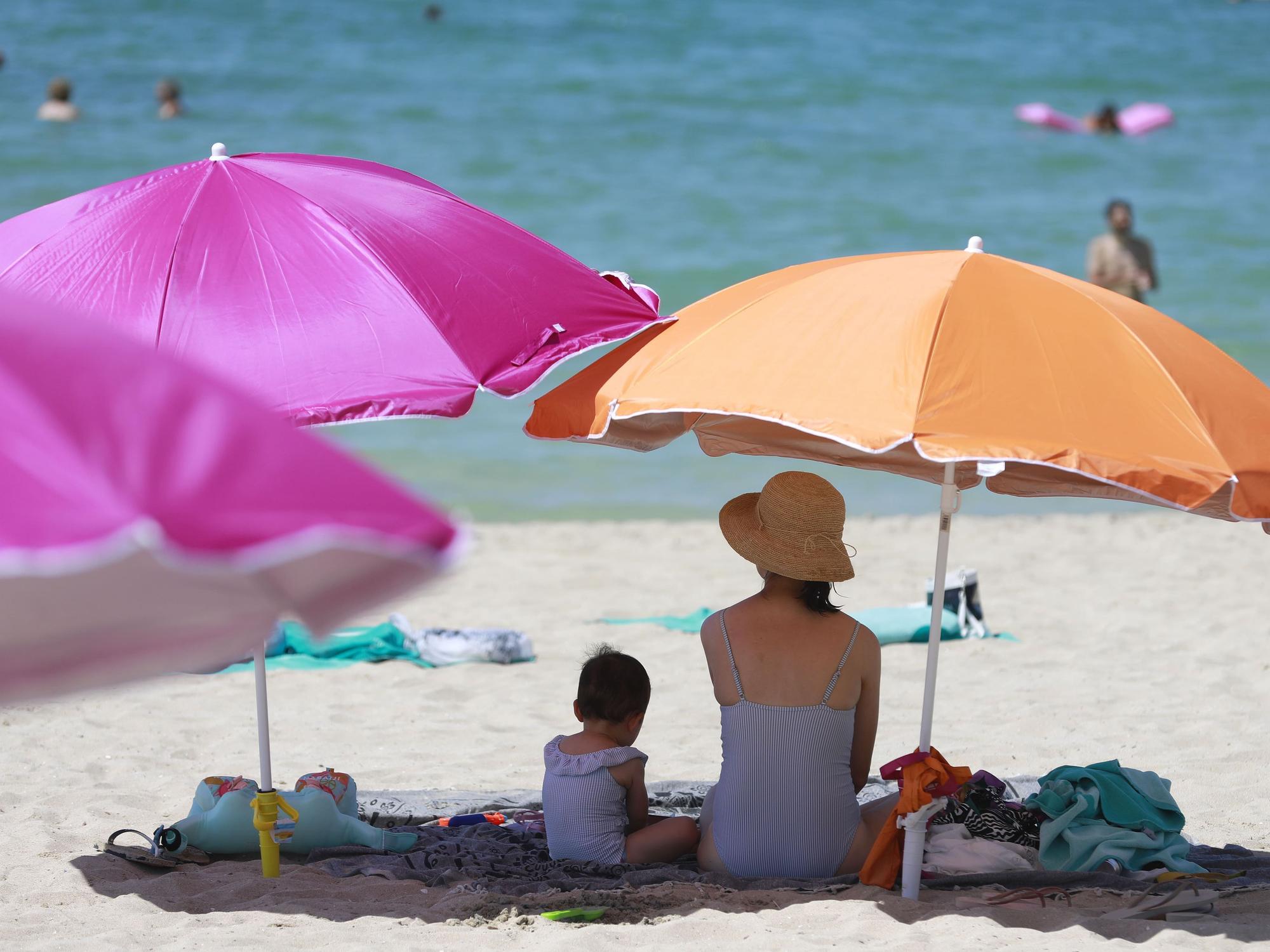 Eine Frau sitzt neben ihrem Kind unter einem Sonnenschirm an einem warmen Sommertag am Strand von Palma de Mallorca im August 2022. Mallorca bleibt das günstigste Reiseziel für Familien im Jahr 2023. Doch einen Haken hat die ganz Sache trotzdem.