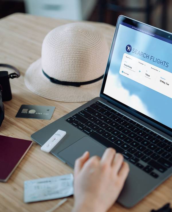 Auf einem Tisch steht ein Laptop, ein Sonnenhut, eine Kamera und ein Reisepass. Auf dem Laptop wird gerade nach Flügen gesucht.