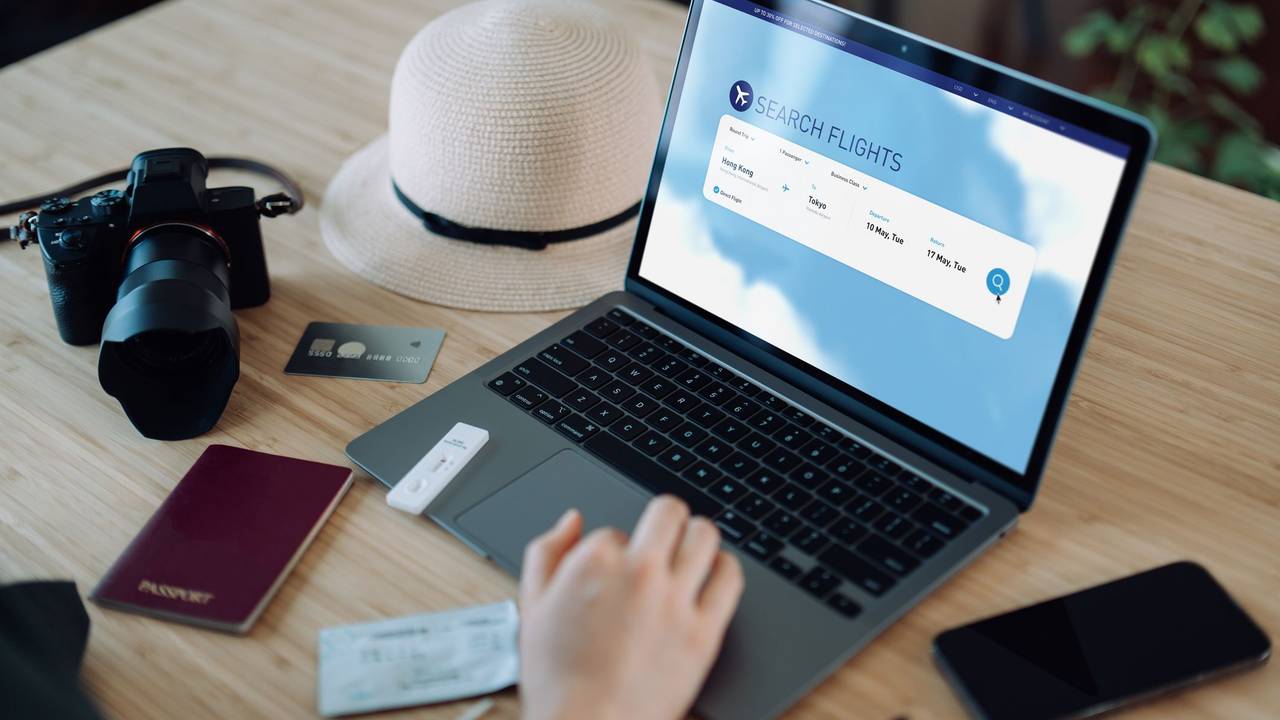 Auf einem Tisch steht ein Laptop, ein Sonnenhut, eine Kamera und ein Reisepass. Auf dem Laptop wird gerade nach Flügen gesucht.