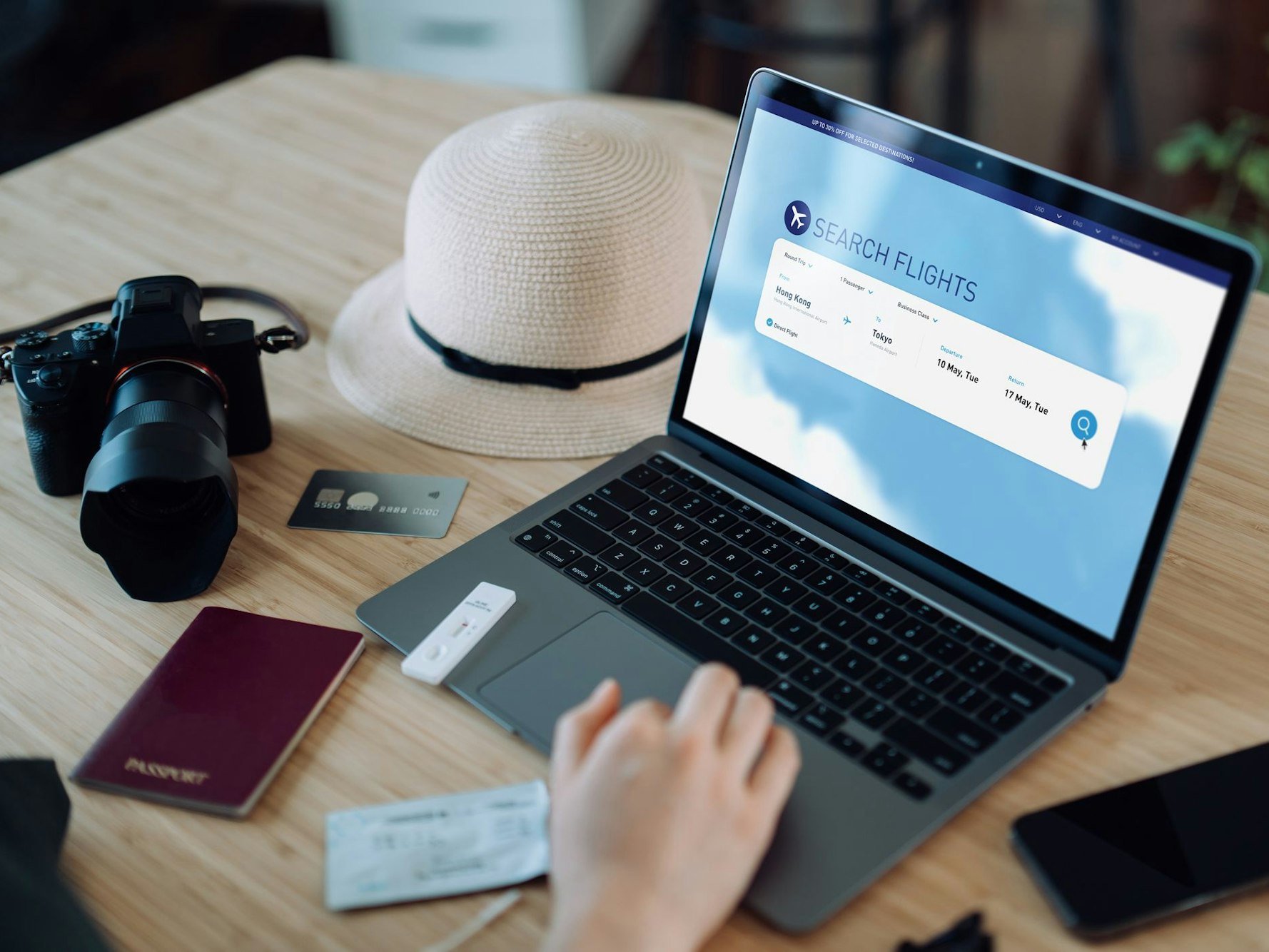Auf einem Tisch steht ein Laptop, ein Sonnenhut, eine Kamera und ein Reisepass. Auf dem Laptop wird gerade nach Flügen gesucht.