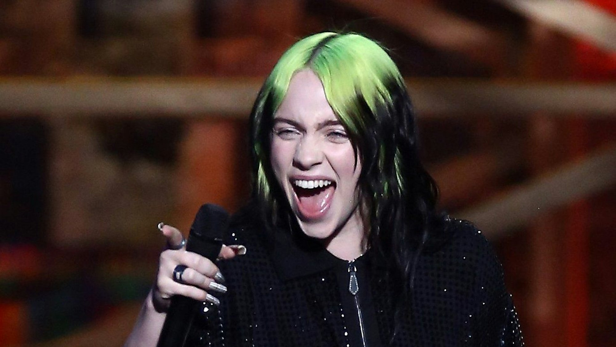 Das Foto aus dem Jahr 2020 zeigt die Sängerin Billie Eilish. Sie hält ein Mikrofon in der rechten Hand, ihr Mund ist geöffnet, die Augen zusammengekniffen. Sie hat grün-schwarze Haare.