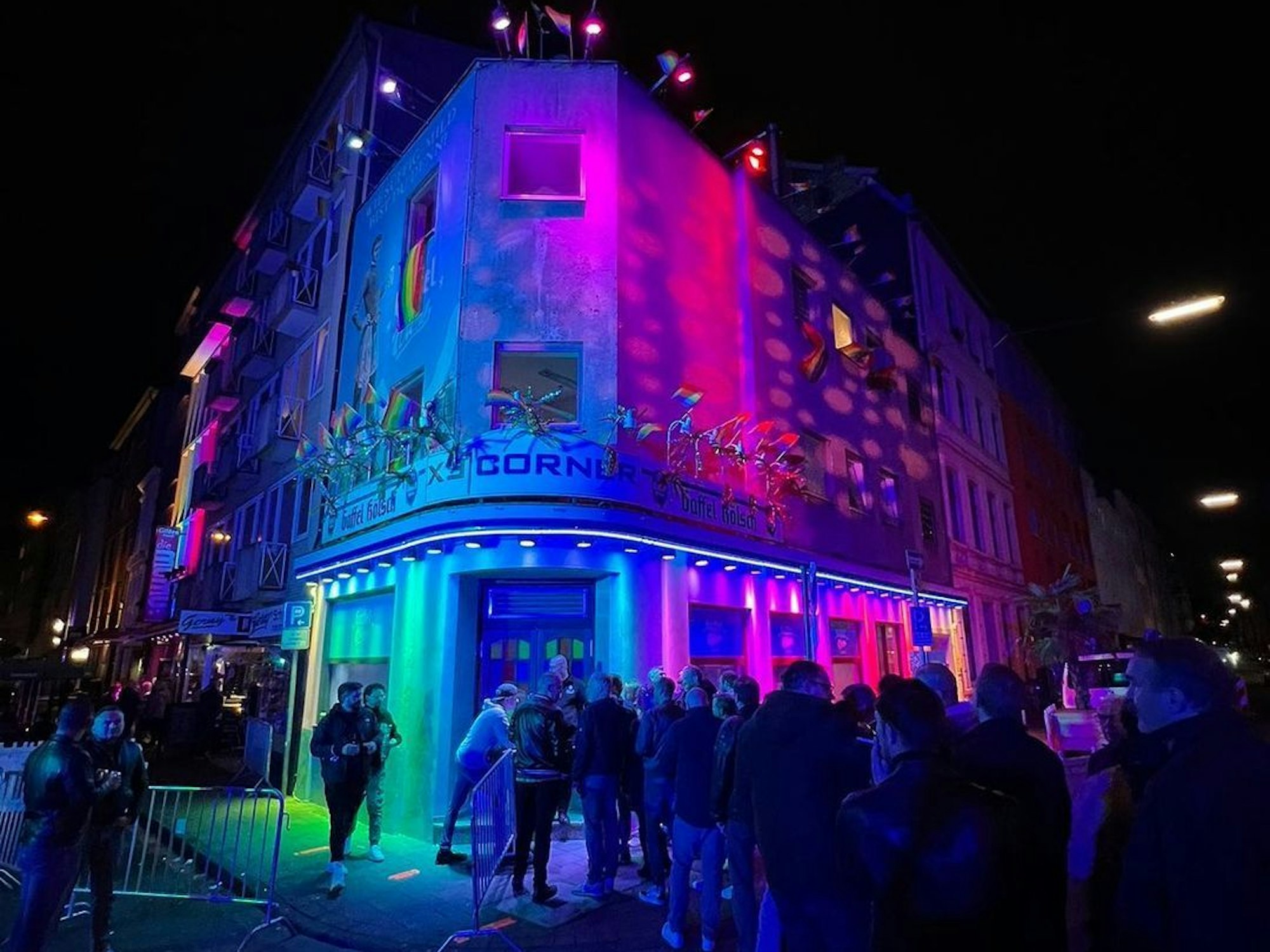Zahlreiche Gäste stehen abends vor der in Regenbogenfarben beleuchteten ExCorner Bar.