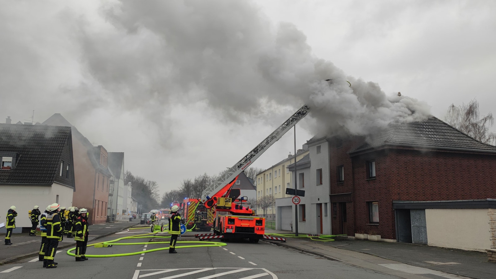 Aus einem Hausdach quillt dichter Rauch, die Feuerwehr hat die Drehleiter ausgefahren. Mehrere Wehrleute stehen auf der Straße.
