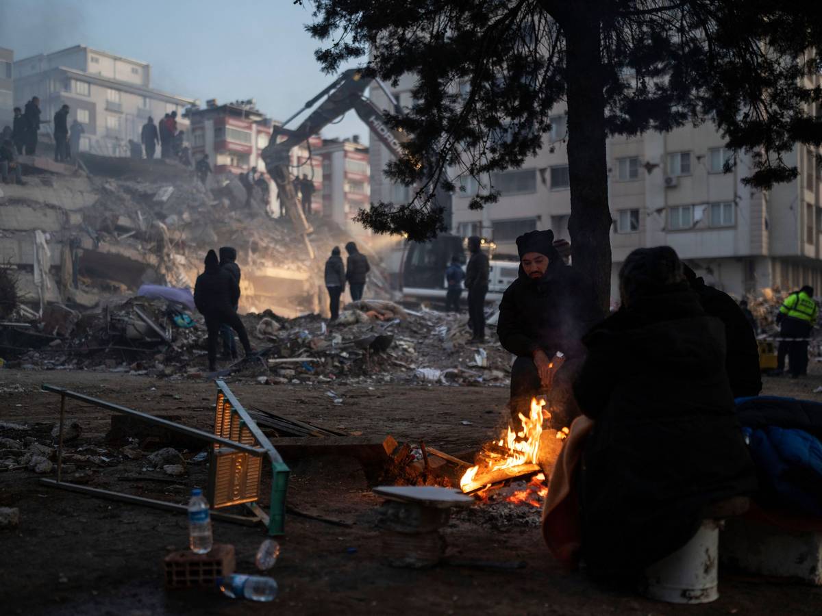 Türkei, Kahramanmaras: Menschen wärmen sich an einem Feuer, während Retter ein durch die Erdbeben zerstörtes Gebäude durchsuchen.