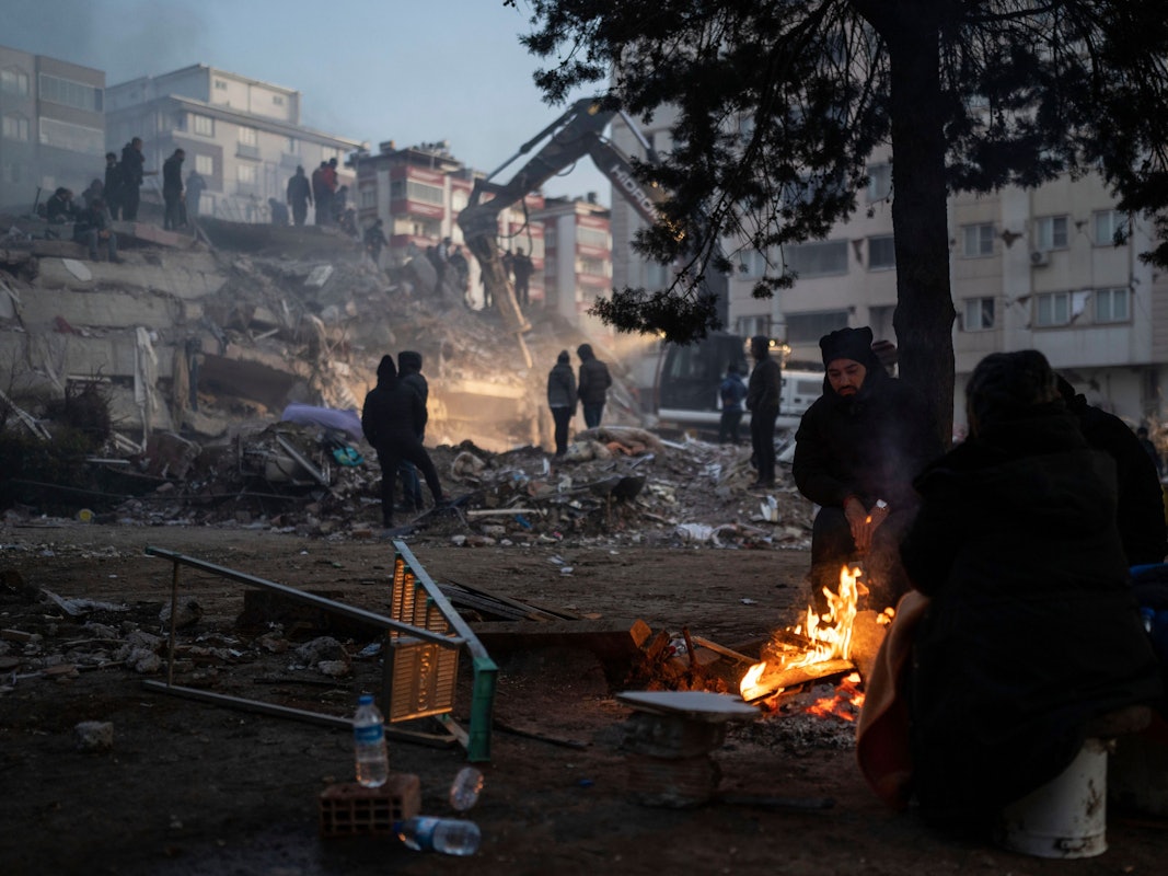 Türkei, Kahramanmaras: Menschen wärmen sich an einem Feuer, während Retter ein durch die Erdbeben zerstörtes Gebäude durchsuchen.