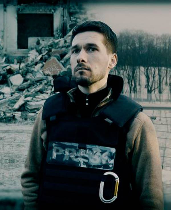 Das Foto zeigt eine Szene aus dem ARD-Film „Ukraine – Krieg im Leben“. Ein Mann mit Schutzweste steht im Vordergrund, hinter ihm sind eine junge Familie und ein zerstörtes Gebäude zu sehen.