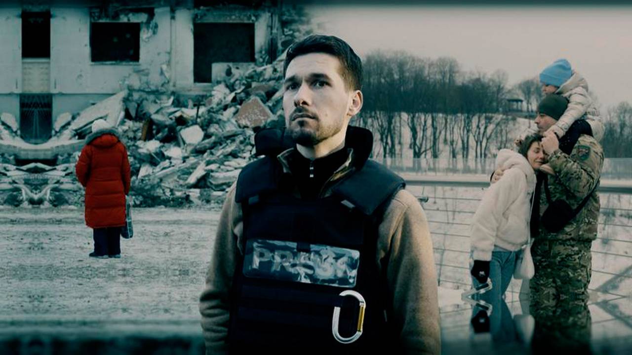 Das Foto zeigt eine Szene aus dem ARD-Film „Ukraine – Krieg im Leben“. Ein Mann mit Schutzweste steht im Vordergrund, hinter ihm sind eine junge Familie und ein zerstörtes Gebäude zu sehen.