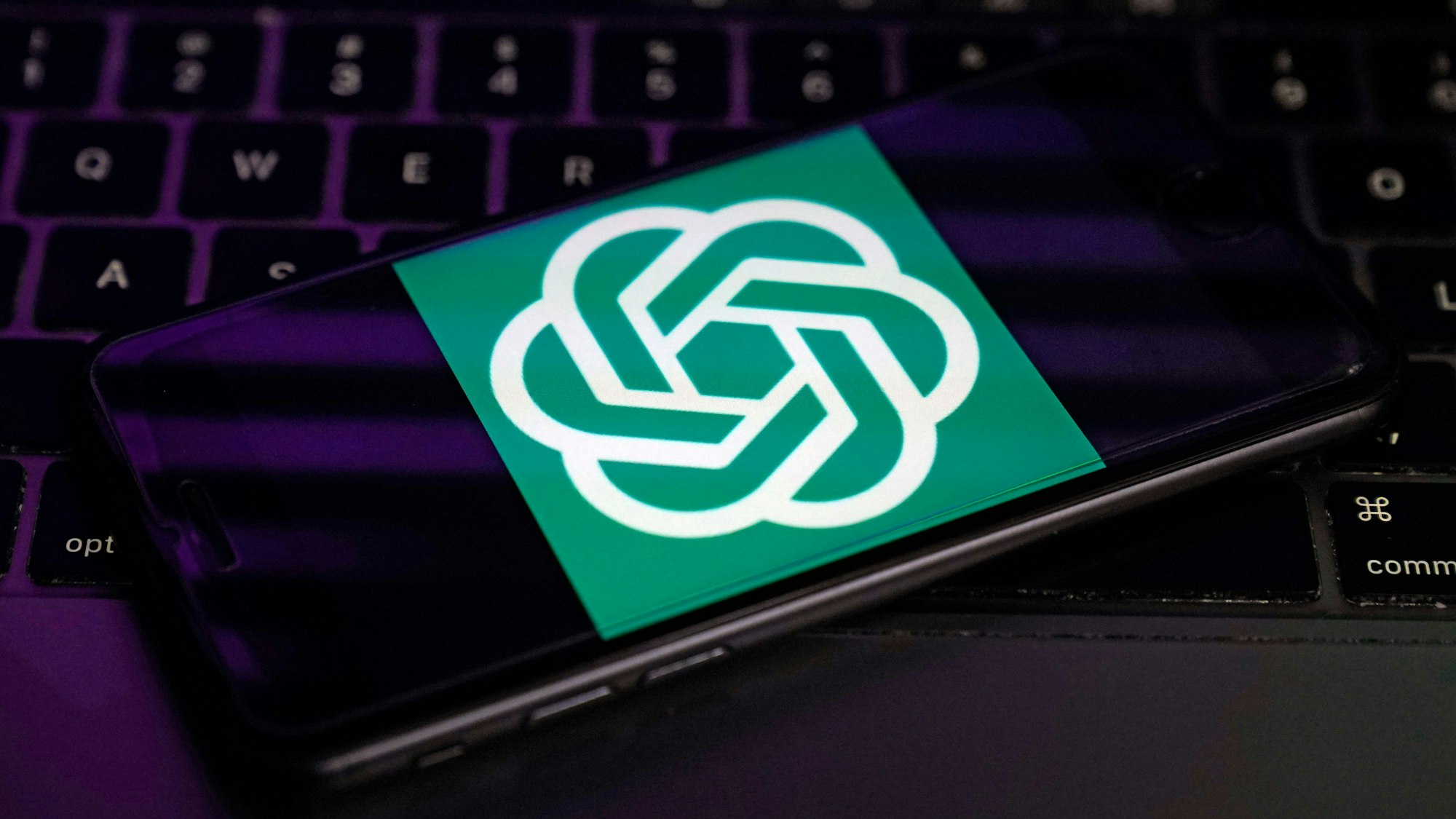 Das undatierte Symbolfoto zeigt ein schwarzes Handy auf einer dunklen Tastatur. Der Handy-Bildschirm zeigt das grün-weiße OpenAI-Logo.