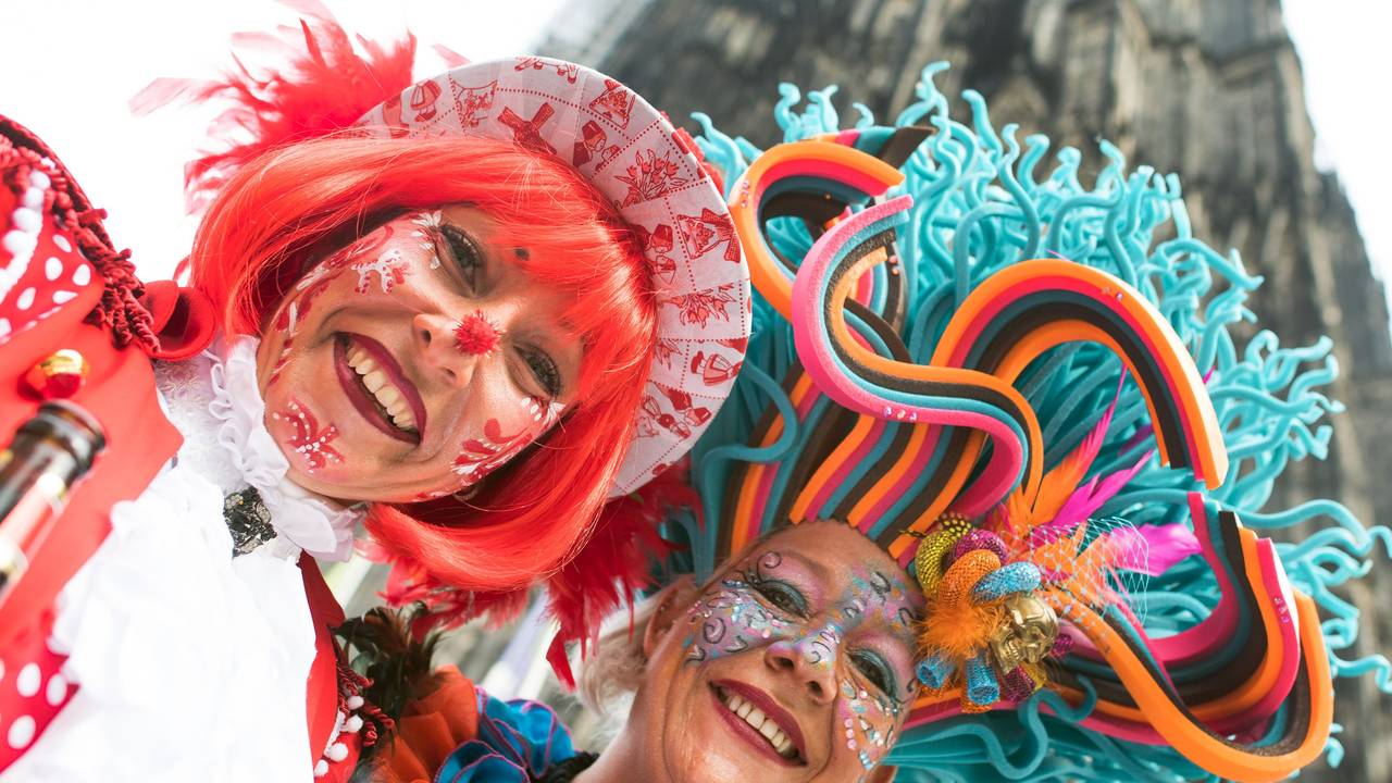 Das Symbolfoto zeigt zwei zu Weiberfastnacht verkleidete Frauen mit bunten Hüten und Karnevalsschminke im Gesicht. Im Hintergrund steht der Kölner Dom.