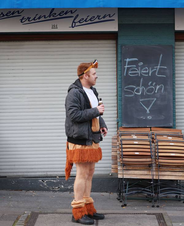 Das Symbolfoto zeigt einen jungen Karnevalisten, der in einem kurzen Löwenkostüm, einer schwarzen Jacke und mit einem Bier in der rechten Hand vor einem geschlossenen Kiosk steht. Auf einer schwarzen Tafel steht „Feiert schön!“.