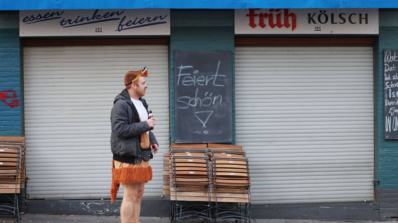 Das Symbolfoto zeigt einen jungen Karnevalisten, der in einem kurzen Löwenkostüm, einer schwarzen Jacke und mit einem Bier in der rechten Hand vor einem geschlossenen Kiosk steht. Auf einer schwarzen Tafel steht „Feiert schön!“.