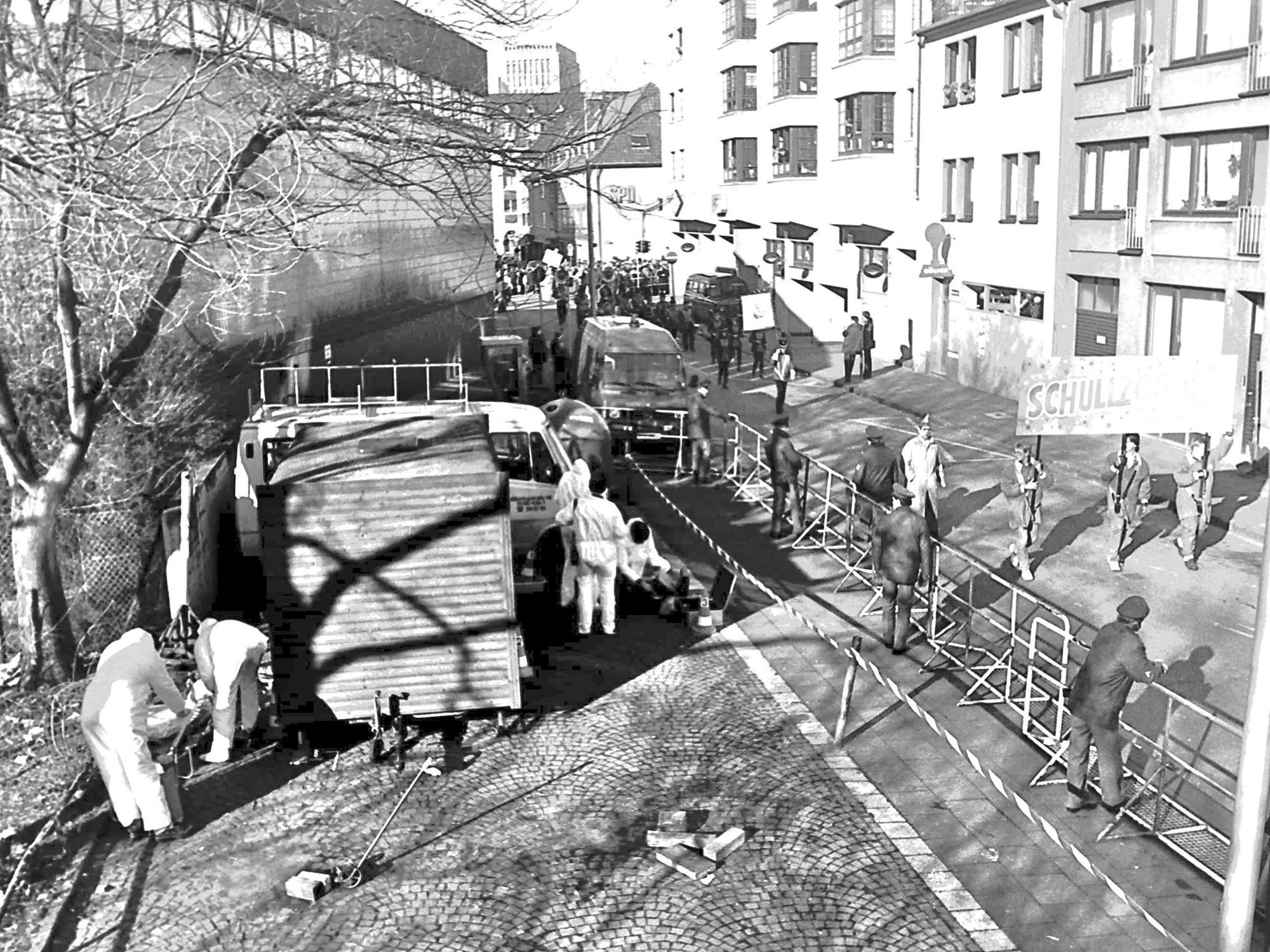 Einsatzkräfte in Schutzanzügen sichern an einem Imbisswagen Spuren, auf der angrenzenden Straße nähert sich ein Karnevalszug.