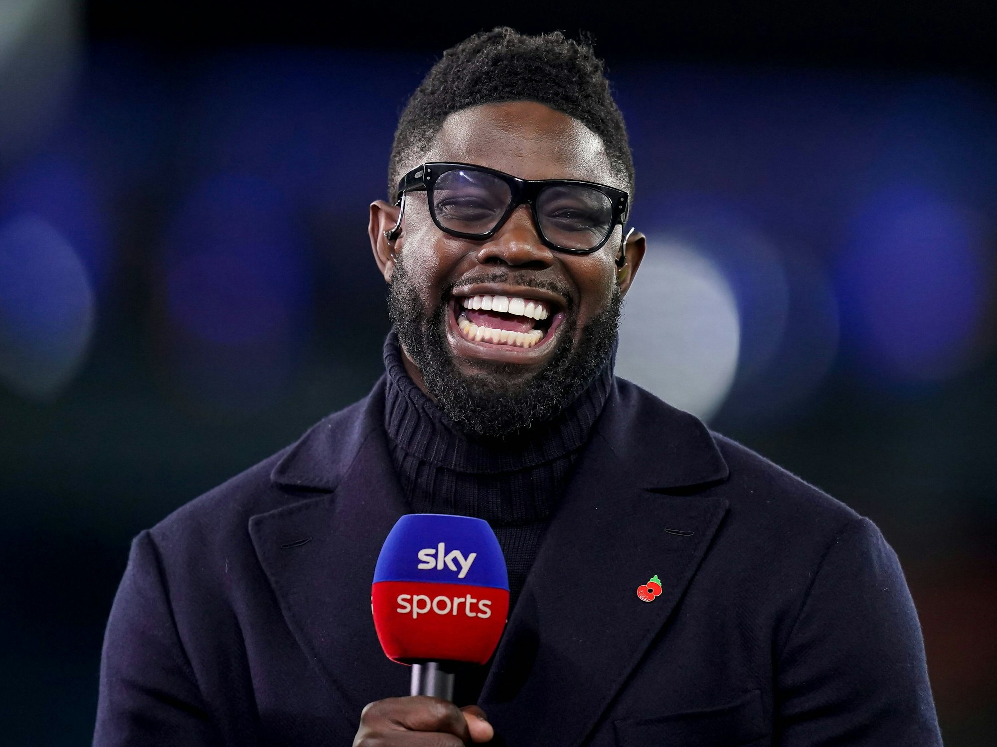 Micah Richards steht mit einem Mikrofon bei einer TV-Übertragung und lacht.