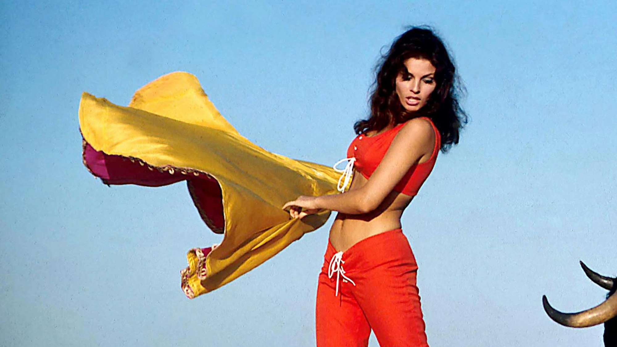 Raquel Welch 1965 bei den Dreharbeiten des Films „Fathom“ in roter Hose und rotem Oberteil mit einem Tuch in der Hand.