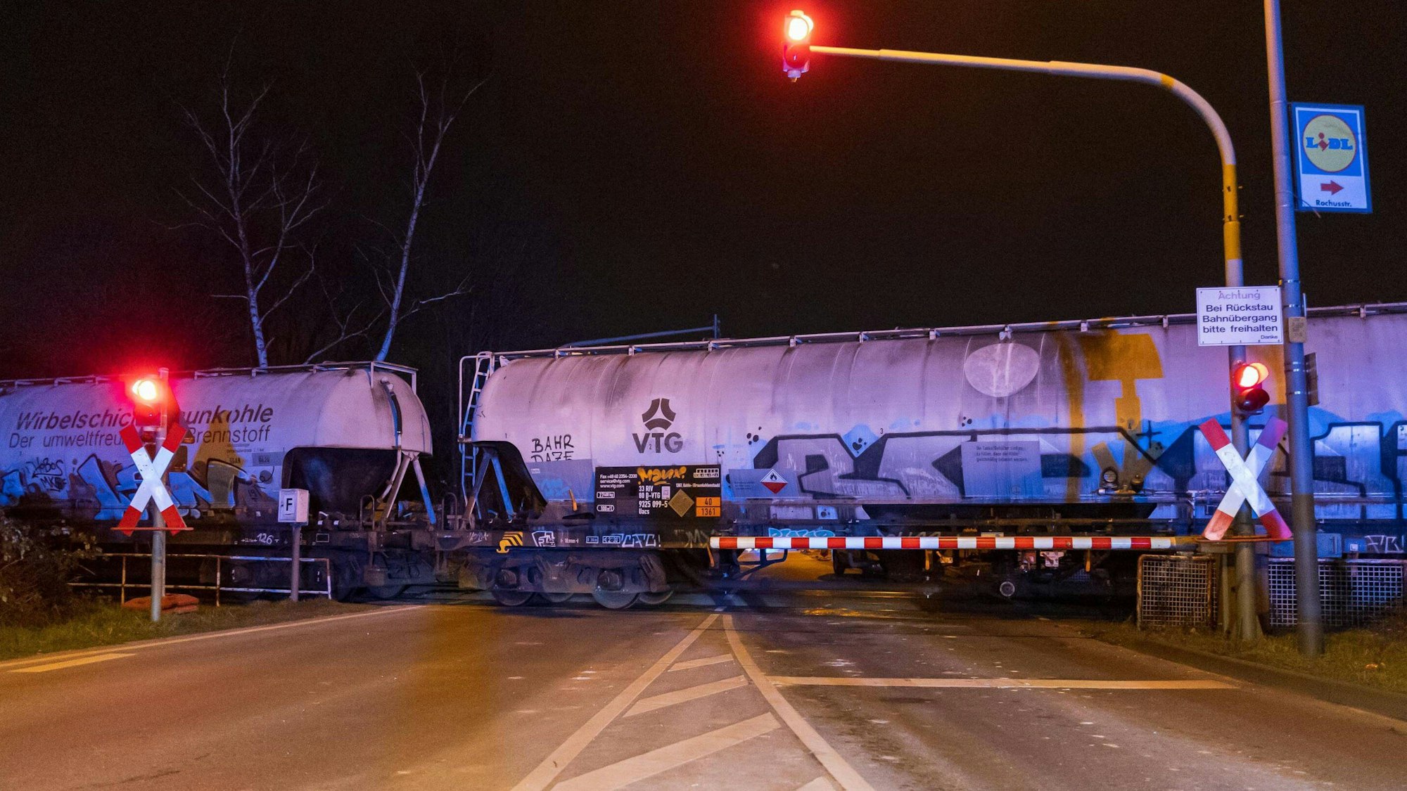 Am Bahnübergang Butzweilerstraße steht ein Güterzug. Die Schranken sind geschlossen, die Ampeln zeigen Rot.