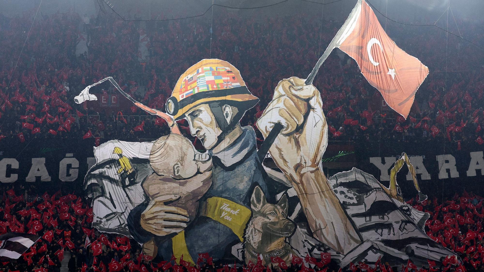 Die Choreografie von Trabzonspor-Fans in der Conference League zeigt einen Helfer nach der Erdbeben-Katastrophe, ein Kind in seinem Arm und das Abbild eines Rettungshundes.