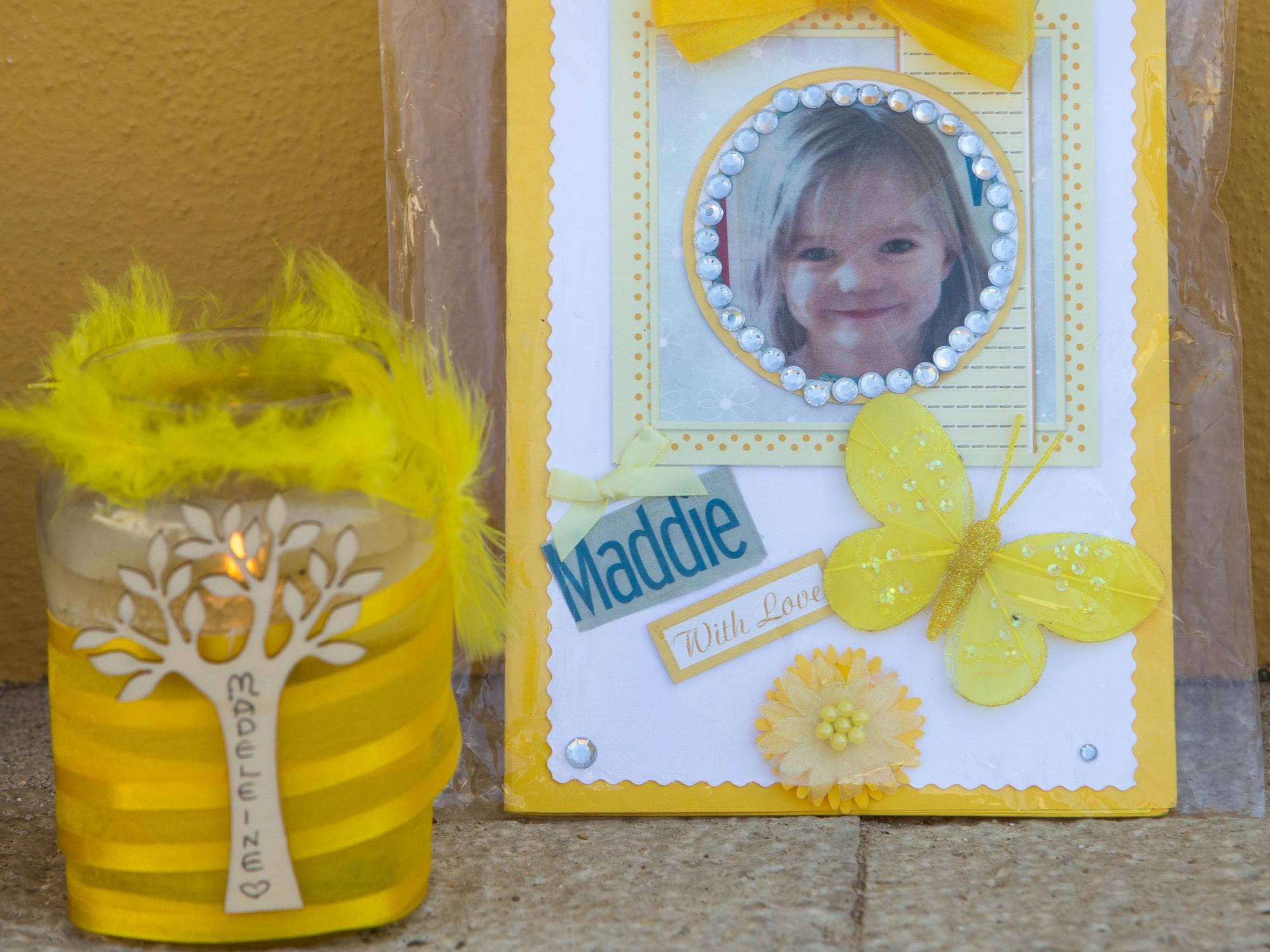 Das Foto von 2017 zeigt eine gelbe Hauswand, vor der eine weiß-gelbe Karte mit dem Foto der vermissten Madeleine McCann und ein gelbes Windlicht aufgestellt sind.