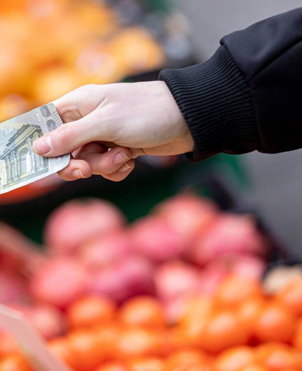 Das Symbolfoto aus dem Jahr 2022 zeigt zwei Hände, die einen 5-Euro-Schein halten. Im Hintergrund sind Kisten mit Obst und Gemüse zu sehen.