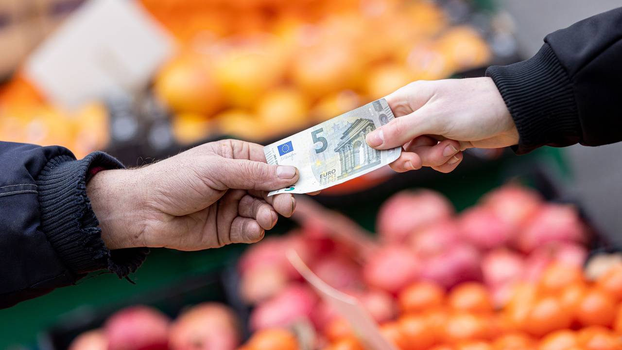 Das Symbolfoto aus dem Jahr 2022 zeigt zwei Hände, die einen 5-Euro-Schein halten. Im Hintergrund sind Kisten mit Obst und Gemüse zu sehen.