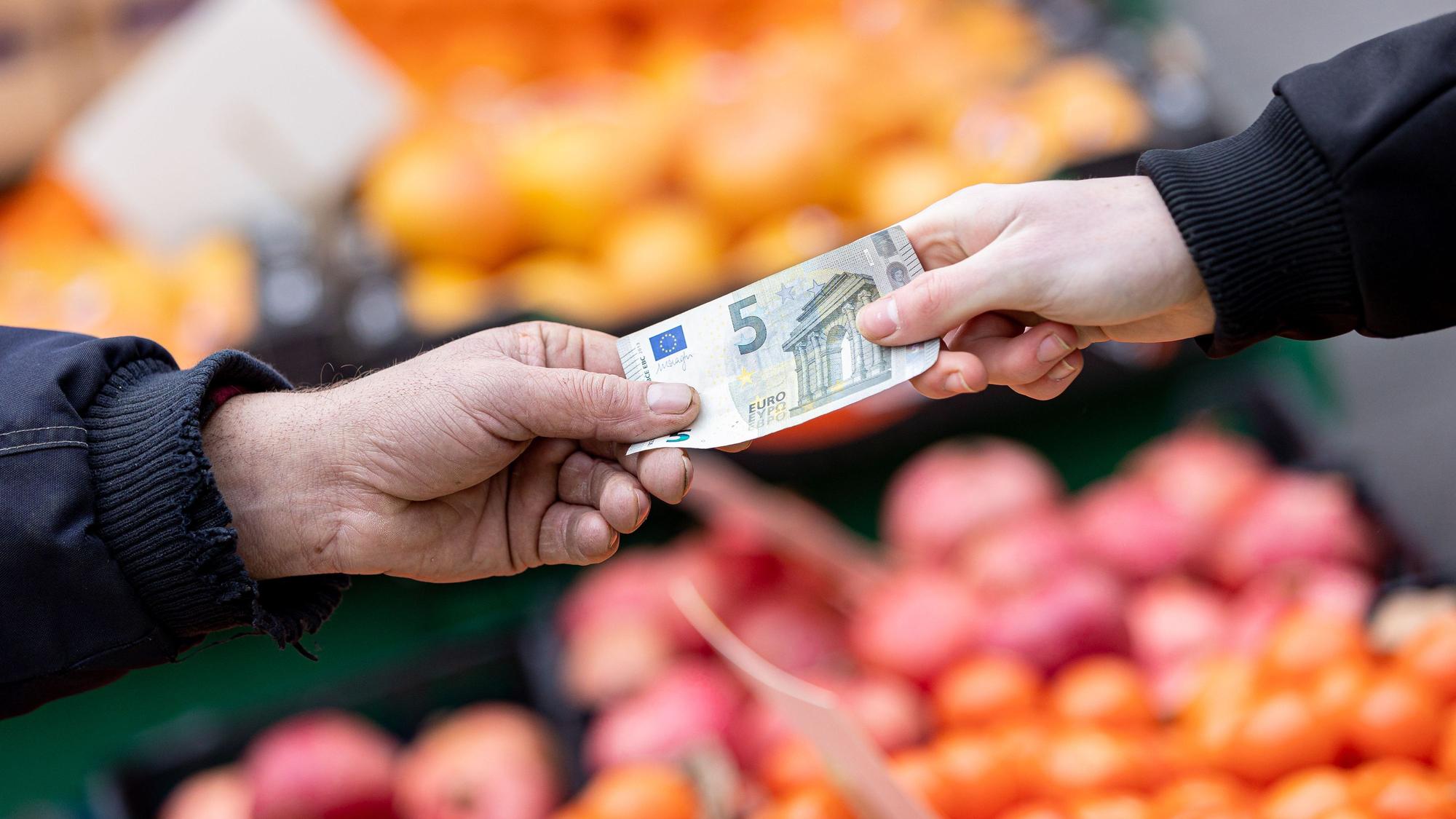 Das Symbolfoto aus dem Jahr 2022 zeigt zwei Hände, die einen 5-Euro-Schein halten. Im Hintergrund sind Kisten mit Obst und Gemüse zu sehen.