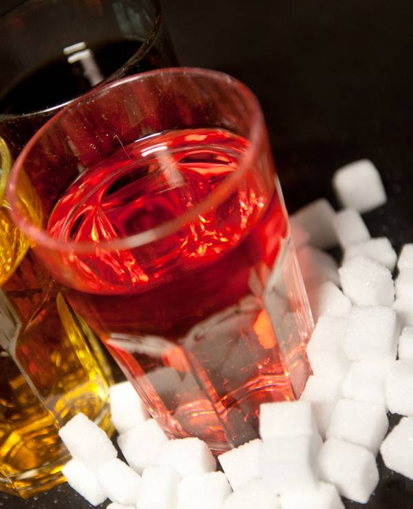 Drei Gläser, gefüllt mit roter Limonade, Cola und einem Energy-Drink stehen neben Zuckerwürfeln auf einem Tisch.