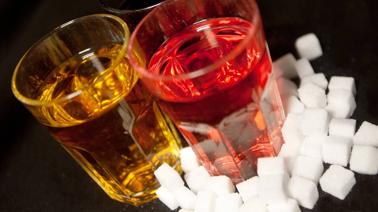Drei Gläser, gefüllt mit roter Limonade, Cola und einem Energy-Drink stehen neben Zuckerwürfeln auf einem Tisch.