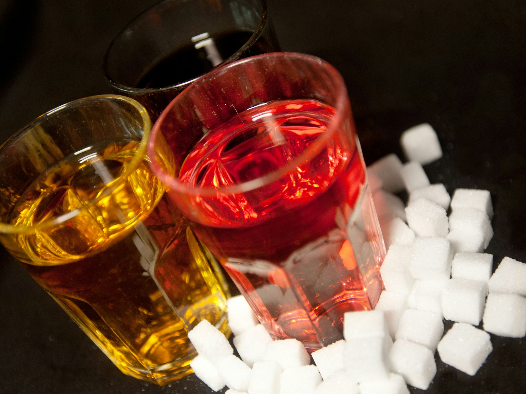 Drei Gläser, gefüllt mit roter Limonade, Cola und einem Energy-Drink stehen neben Zuckerwürfeln auf einem Tisch.