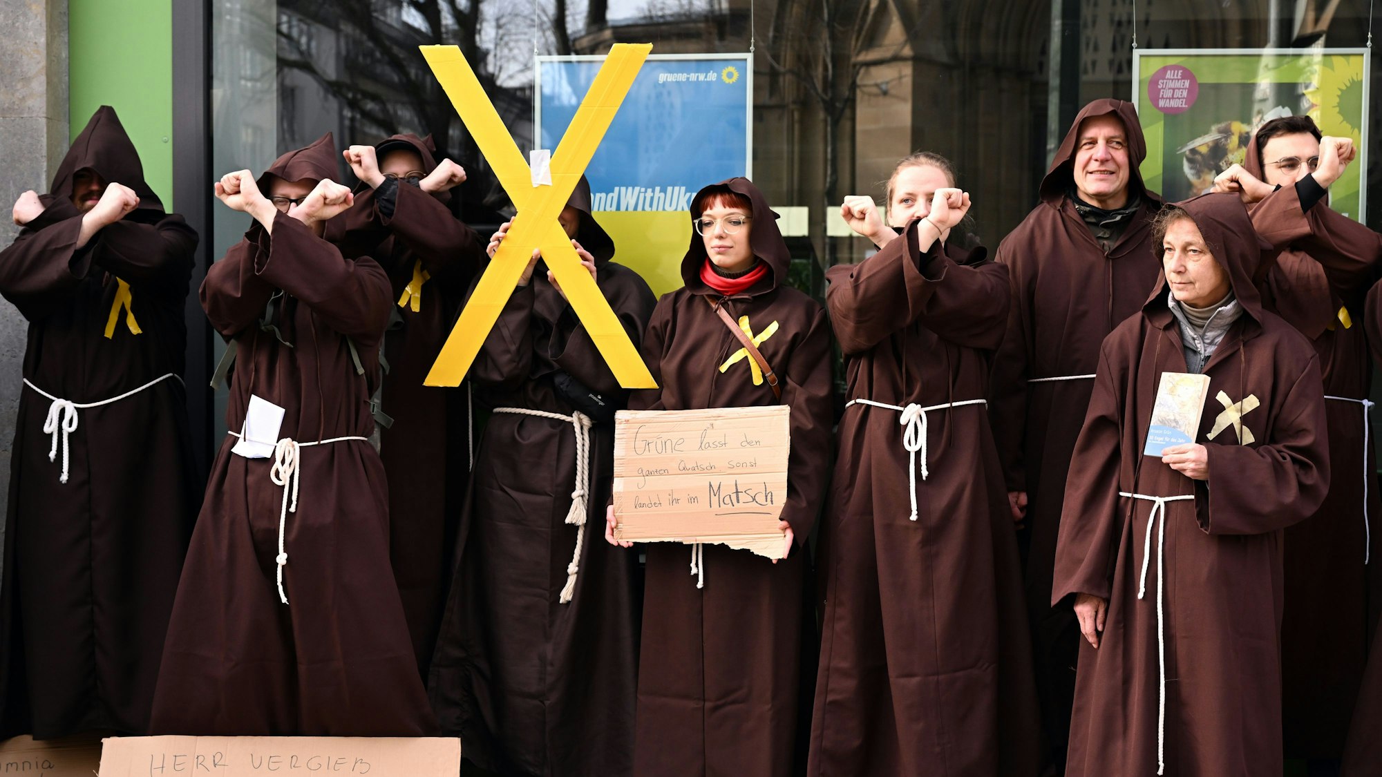Das Foto vom 22. Februar 2023 zeigt als Mönche verkleidete Menschen vor der Grünen-Parteizentrale in Düsseldorf. Eine Person hält ein gelbes X in die Höhe.