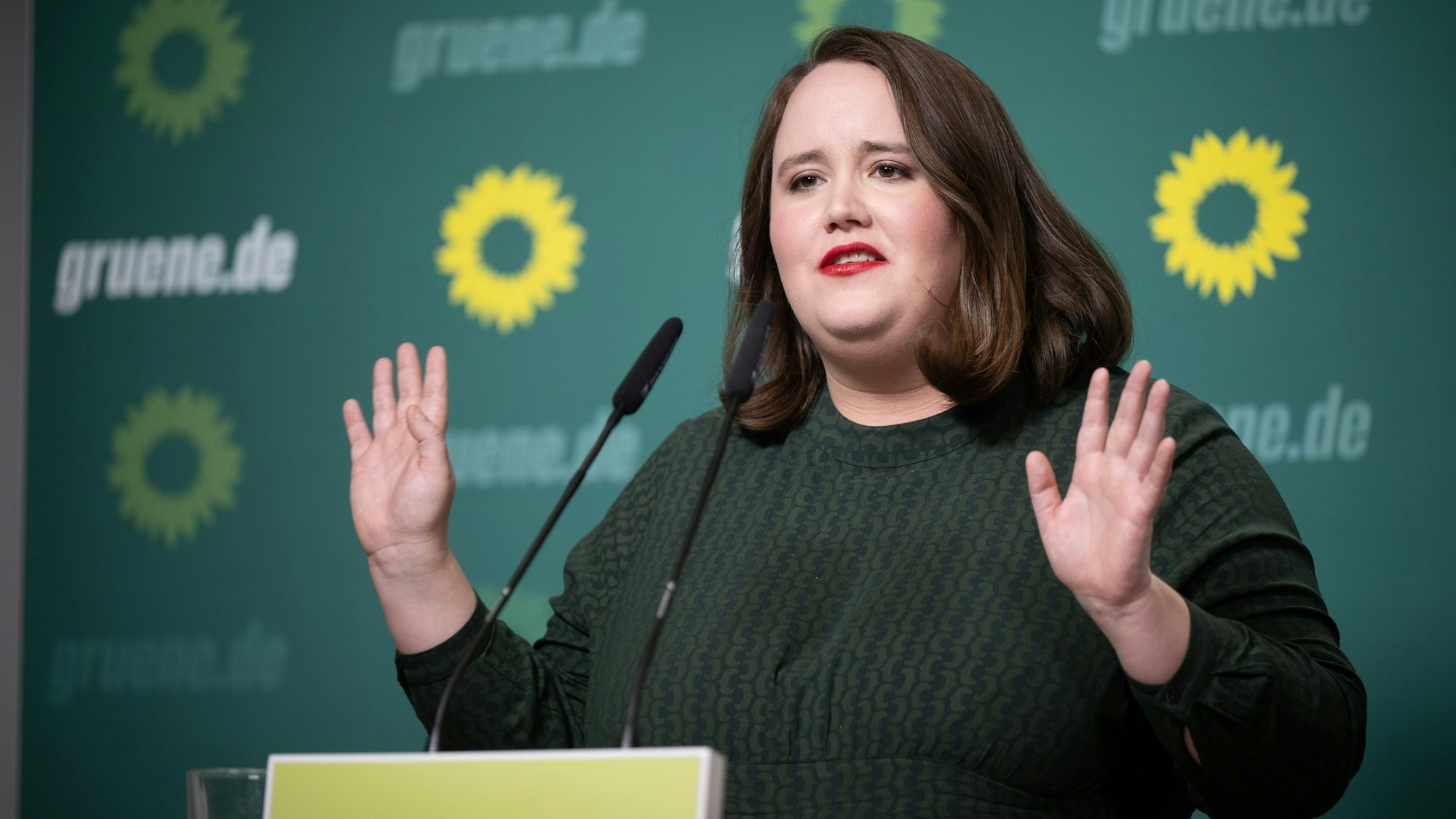 Das Foto zeigt die Grünen-Vorsitzende Ricarda Lang. Sie steht vor einem grünen Hintergrund, trägt einen grünen Pullover, vor ihr ist ein Mikrofon zu sehen. Lang hebt beide Hände.