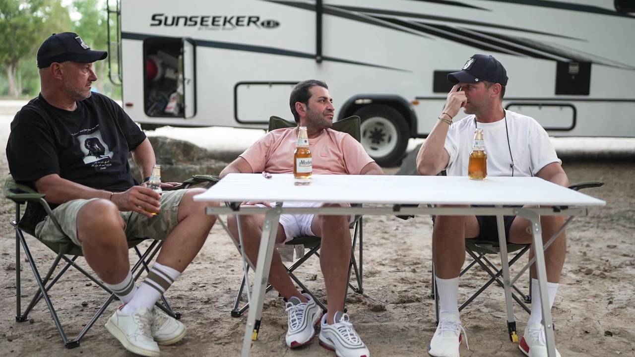 Frank Rosin, Ali Güngörmüş und Alexander Kumptner sitzen an einem Campingtisch vor einem Wohnmobil.
