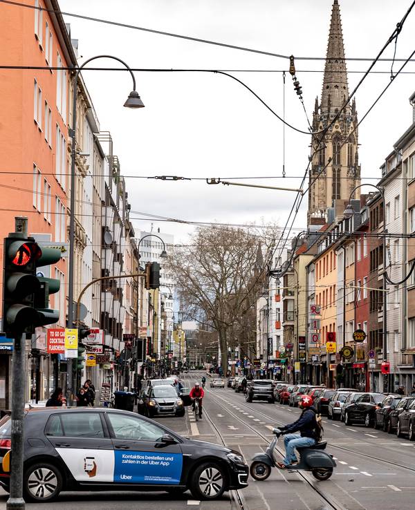 Blick in die Zülpicher Straße in Köln. Neben Autos und einer Vespa ist im Hintergrund ein Kirchturm zu sehen.
