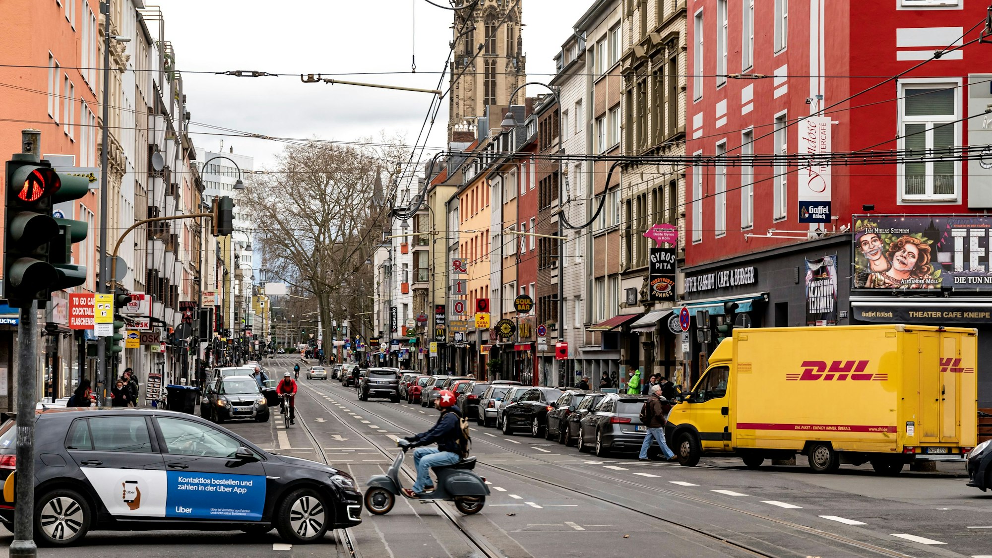 Blick in die Zülpicher Straße in Köln. Neben Autos und einer Vespa ist im Hintergrund ein Kirchturm zu sehen.