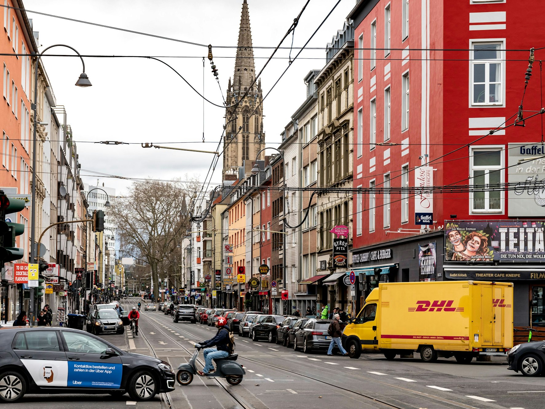 Blick in die Zülpicher Straße in Köln. Neben Autos und einer Vespa ist im Hintergrund ein Kirchturm zu sehen.