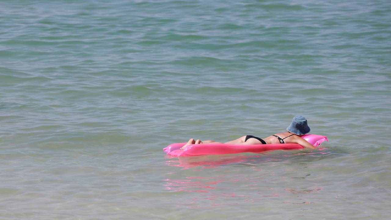 Das Symbolfoto zeigt eine Frau im Bikini. Sie trägt einen blauen Hut und liegt bäuchlings auf einer pinken Luftmatratze, die auf dem Meer treibt.