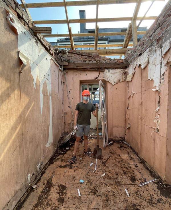 Ein junger Mann steht inmitten eines zerbombten Hauses. Er trägt einen Schutzhelm und eine Gesichtsmaske. In seiner Hand hält er eine Schaufel.