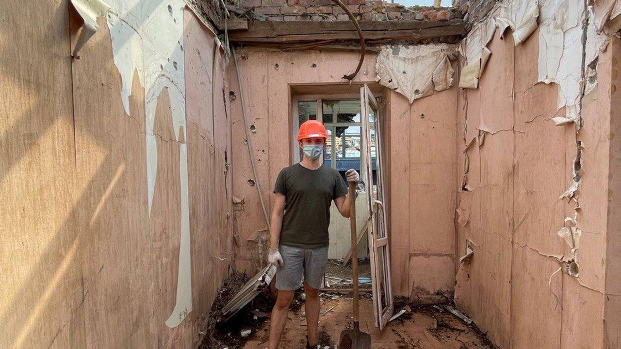 Ein junger Mann steht inmitten eines zerbombten Hauses. Er trägt einen Schutzhelm und eine Gesichtsmaske. In seiner Hand hält er eine Schaufel.