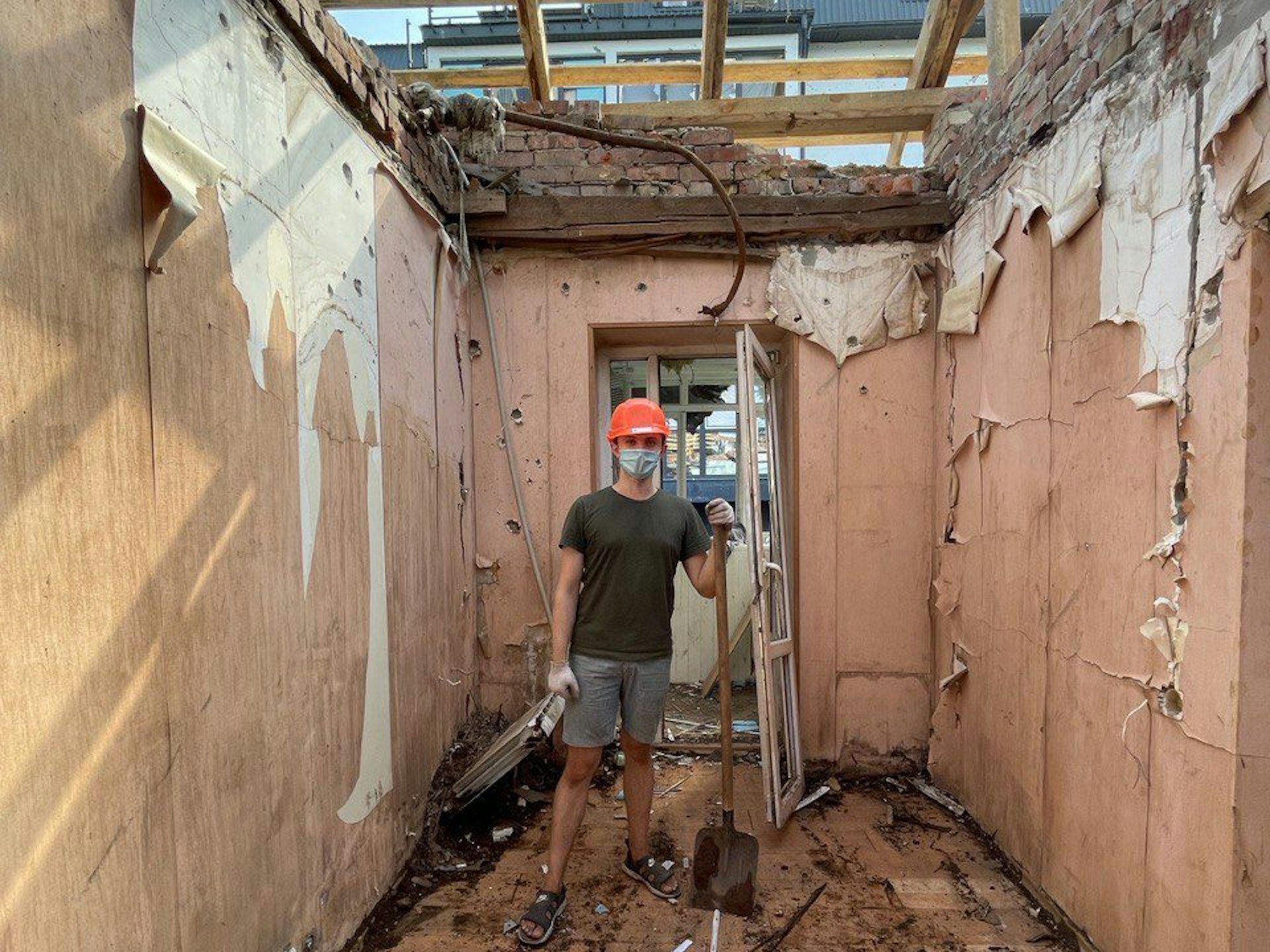 Ein junger Mann steht inmitten eines zerbombten Hauses. Er trägt einen Schutzhelm und eine Gesichtsmaske. In seiner Hand hält er eine Schaufel.