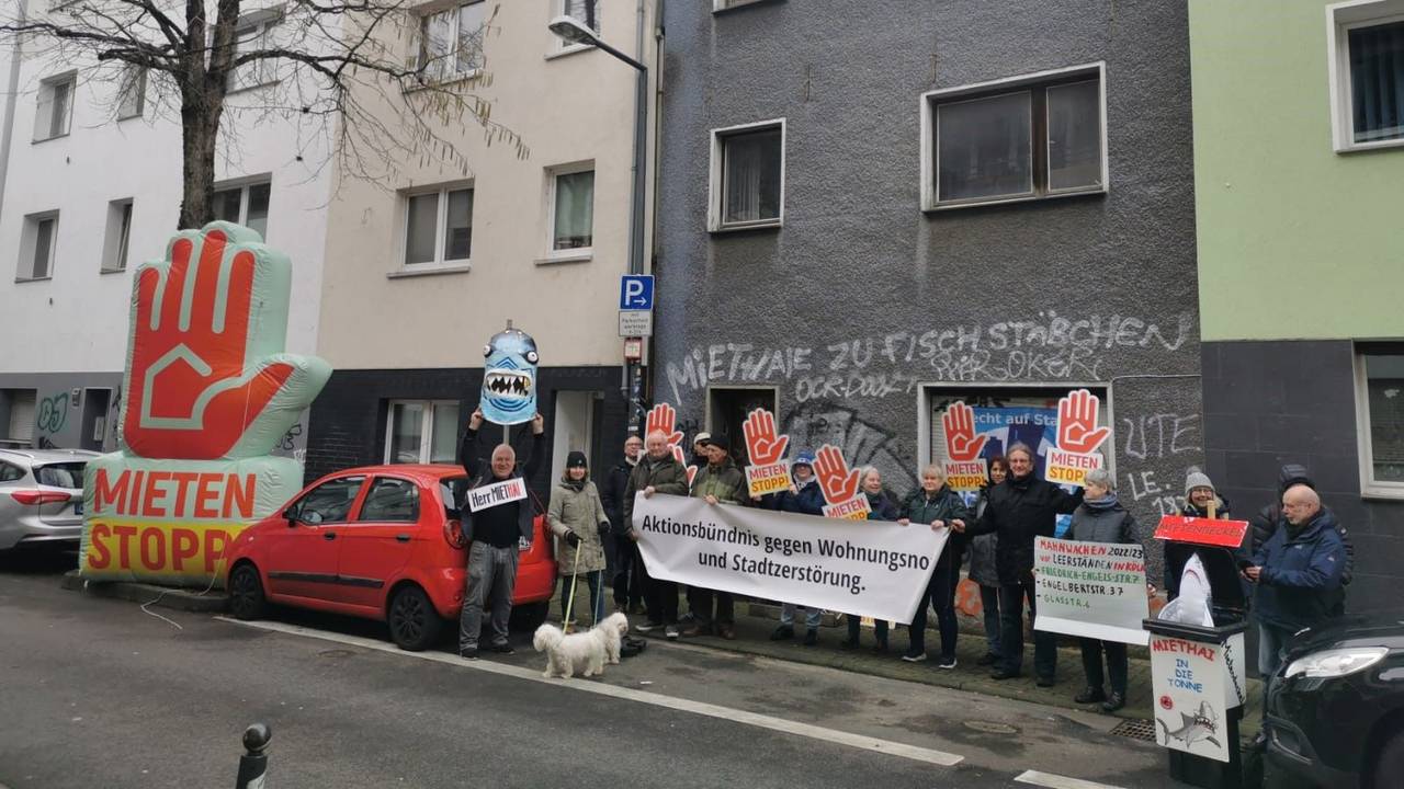 Männer und Frauen haben sich vor einem leerstehenden Haus positioniert. Einige halten ein Transparent mit der Aufschrift „Aktionsbündnis gegen Wohnungsnot und Stadtzerstörung“, andere Plakate mit der Forderung „Mieten Stopp“.