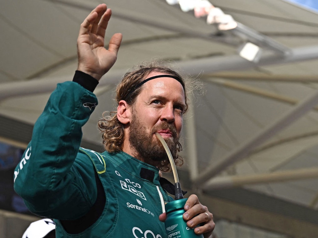 Sebastian Vettel trinkt und winkt dabei im Rennanzug von Aston Martin den Fans zu.