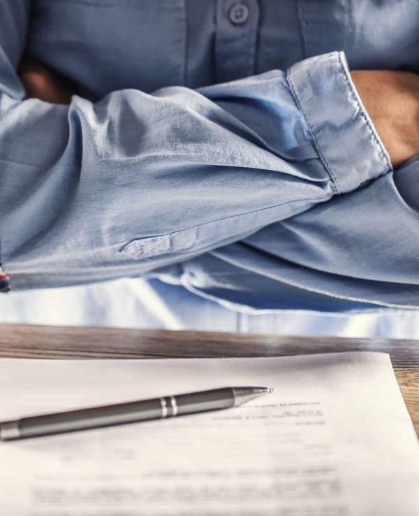 Ein Mann kreuzt die Arme vor der Brust, vor ihm auf dem Tisch liegt ein Stapel Unterlagen.