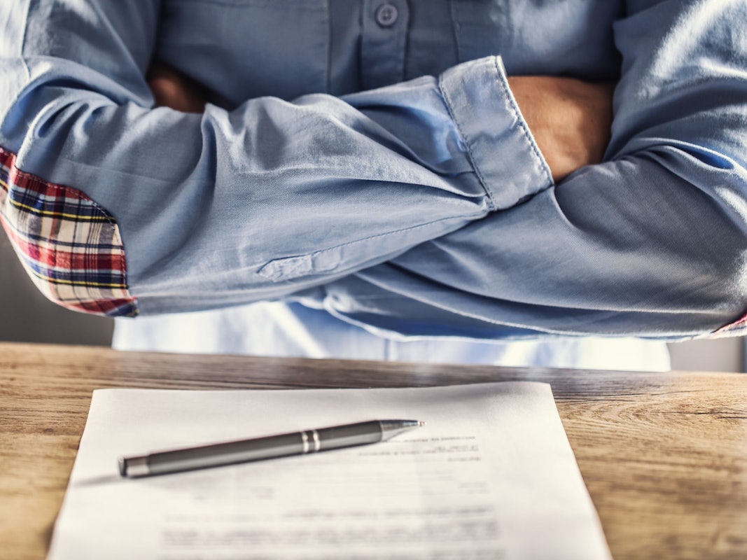Ein Mann kreuzt die Arme vor der Brust, vor ihm auf dem Tisch liegt ein Stapel Unterlagen.