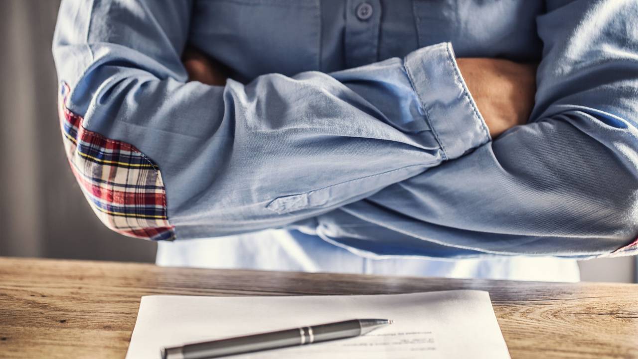 Ein Mann kreuzt die Arme vor der Brust, vor ihm auf dem Tisch liegt ein Stapel Unterlagen.