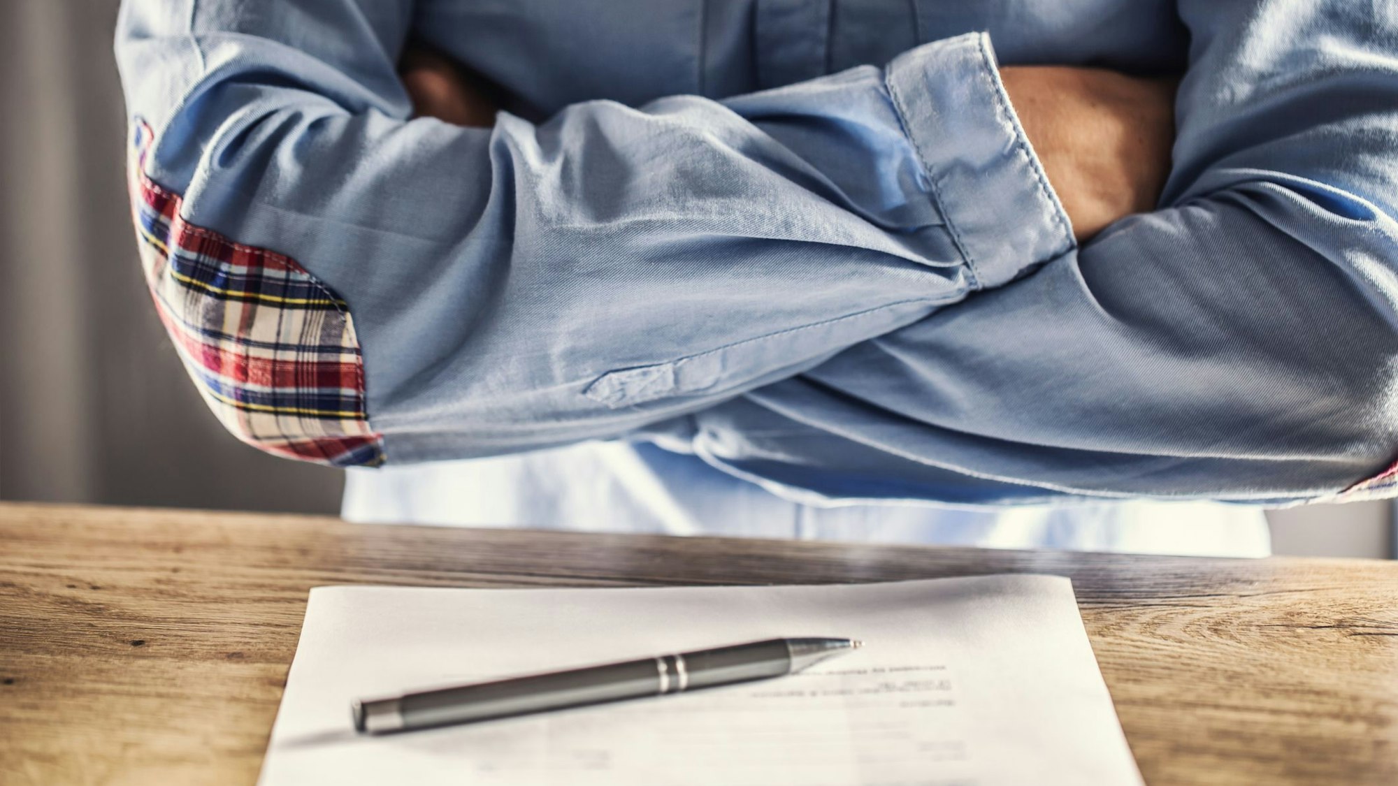 Ein Mann kreuzt die Arme vor der Brust, vor ihm auf dem Tisch liegt ein Stapel Unterlagen.