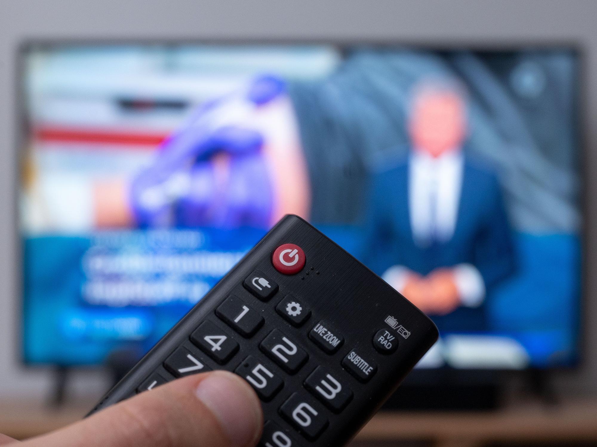 Ein Fernseher, auf dem Tagesschau läuft. Im Vordergrund: ein Finger auf einer Fernbedienung.