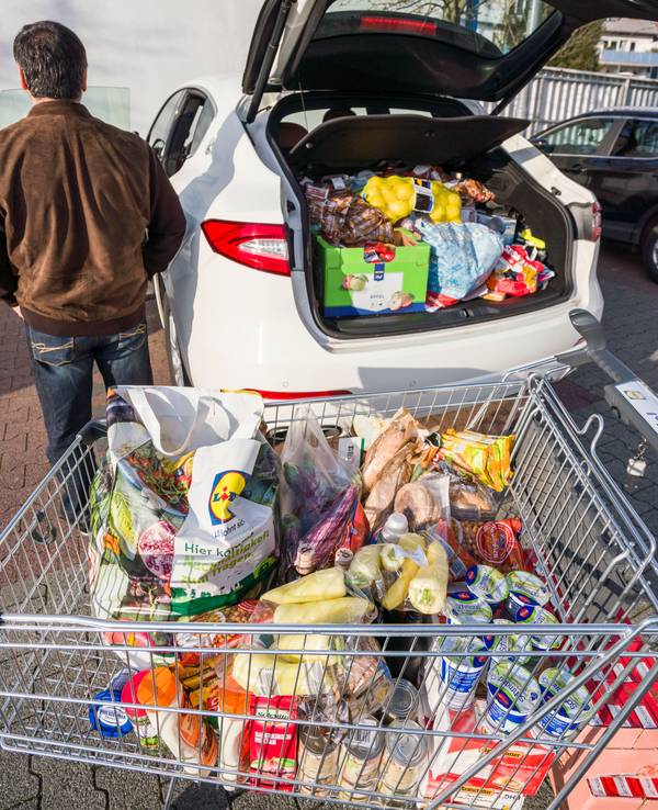 Ein Mann steht nach seinem Einkauf bei einem Discounter neben seinem vollen Einkaufswagen und dem bereits mit Lebensmitteln gut gefüllten Kofferraum seines Autos.