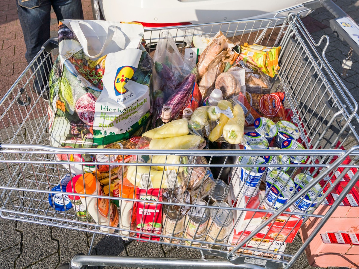 Ein Mann steht nach seinem Einkauf bei einem Discounter neben seinem vollen Einkaufswagen und dem bereits mit Lebensmitteln gut gefüllten Kofferraum seines Autos.