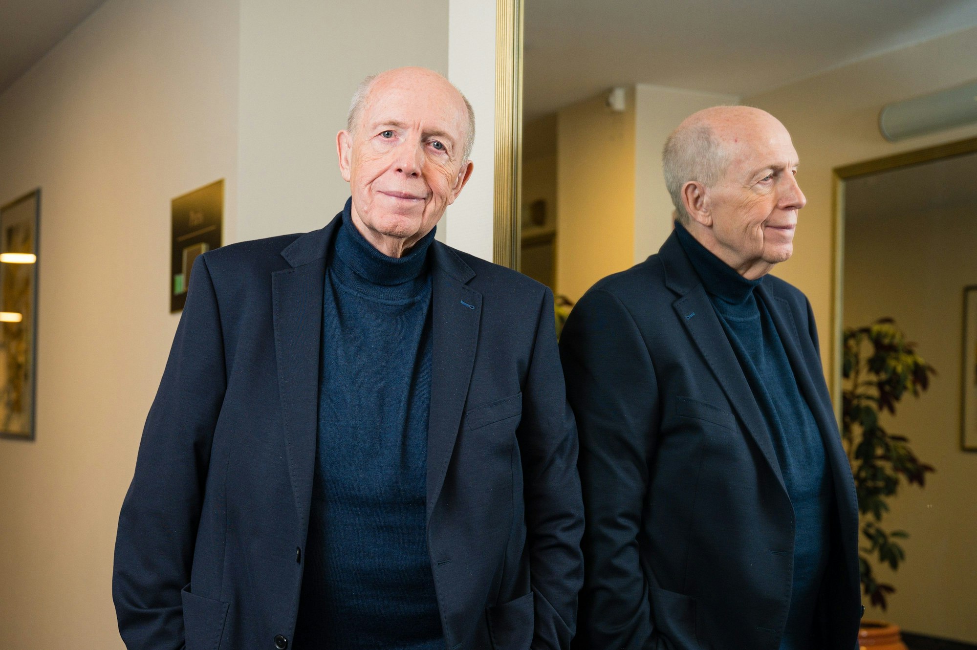 Reiner Calmund steht in einem dunklen Anzug neben einem Spiegel und blickt lächelnd in die Kamera.