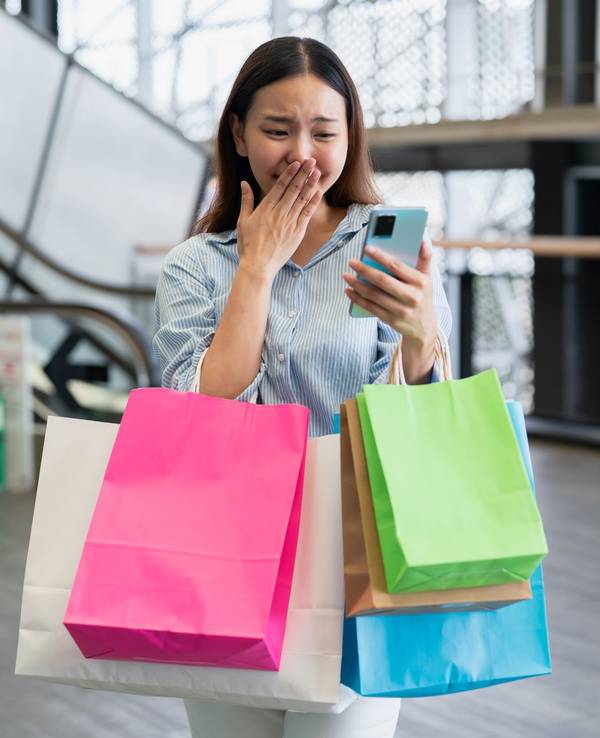 Eine Frau, bepackt mit bunten Tüten, schaut aufgeregt auf ihr Handy.