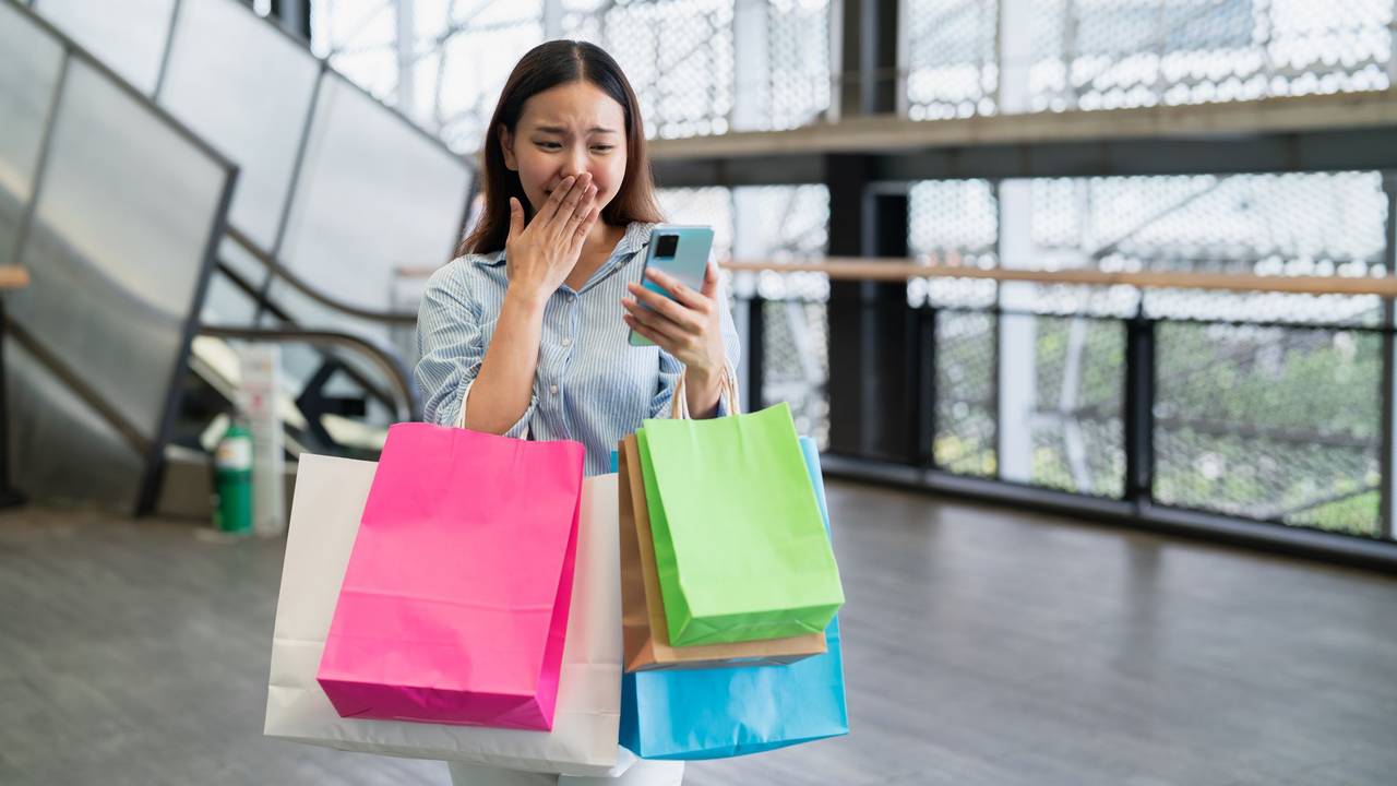 Eine Frau, bepackt mit bunten Tüten, schaut aufgeregt auf ihr Handy.