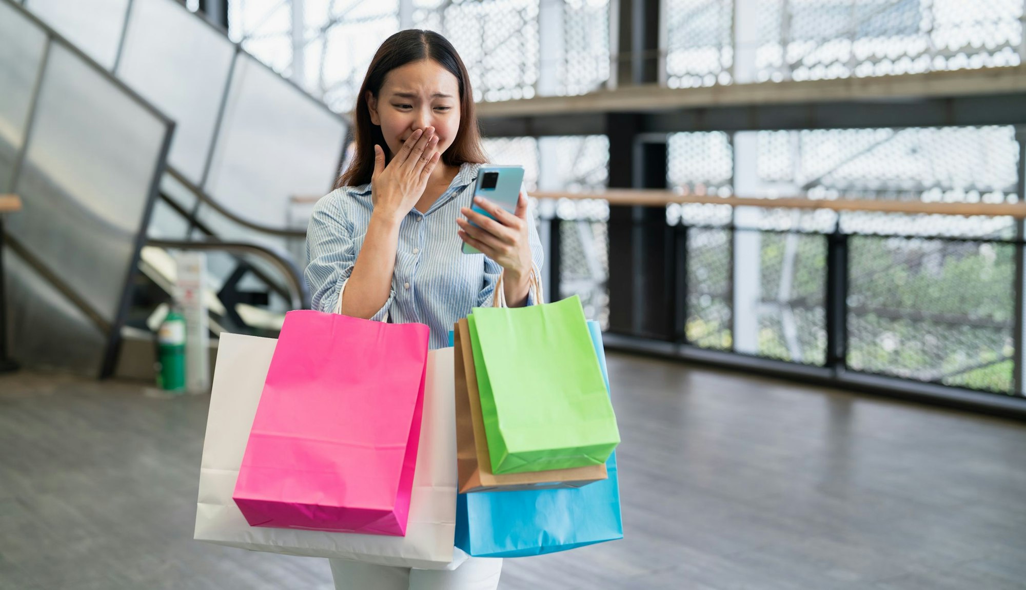 Eine Frau, bepackt mit bunten Tüten, schaut aufgeregt auf ihr Handy.